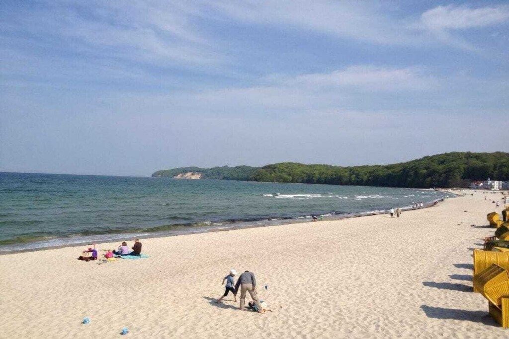 Une plage où l’on voit des gens se prélasser au soleil et se promener, un ciel dégagé et, au loin, une colline boisée.