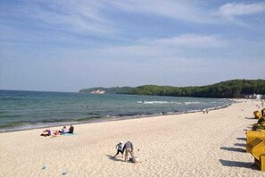 Une plage où l’on voit des gens se prélasser au soleil et se promener, un ciel dégagé et, au loin, une colline boisée.
