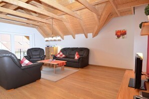 Un vaste salon doté d’un haut plafond et de poutres apparentes, avec deux canapés en cuir noir ornés de coussins à carreaux rouges et blancs, une table basse en bois et un téléviseur posé sur un meuble en bois.