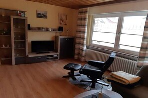 A living room with a television, a black office chair, a sofa, a radiator, and a window with curtains. - Holiday apartment with 2 bedrooms (Rinteln)