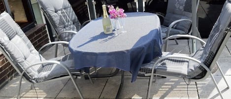 An outdoor patio with a round table covered by a blue tablecloth, surrounded by metal chairs.