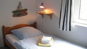 A single bed with a wooden headboard, a bedside lamp, a small shelf with a lamp, and a window with curtains. - Holiday home in Fehmarn with swimming pool (Fehmarn)
