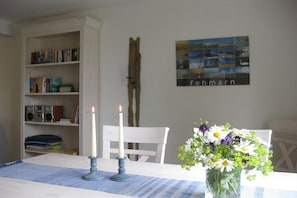 Een kamer met een witte tafel, twee kaarsen, een bloemenvaas, een boekenkast en een aan de muur hangend schilderij.