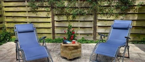 Zwei blaue Lounge-Stühle mit Metallgestellen, eine Holz-Pflanzkiste mit Blumen und einer roten Tasse, sowie ein Holzzaun mit Kletterpflanzen.