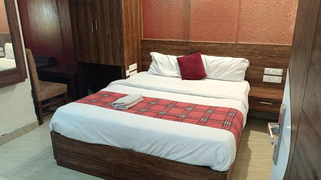 Ein Doppelbett mit einer rot-weißen Karodecke, weißen Kissen und einem roten Zierkissen. Eine Holzgarderobe befindet sich links, und ein kleiner Tisch mit einem Stuhl steht daneben.