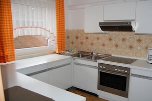 Cuina amb armaris blancs, forn d'acer inoxidable, rentapelats i finestra amb cortines taronges.