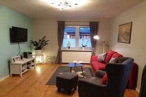 A living room with a red sofa, a black armchair, a small table, a TV on the wall, and a window with curtains. - Holiday apartment with 1 bedroom (Niendorf  a.Fehmarn)