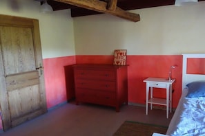 Ein Zimmer mit einer Holztür, einem roten Schminktisch, einem weißen Nachttisch und einem Bett mit einem blauen Kissen.