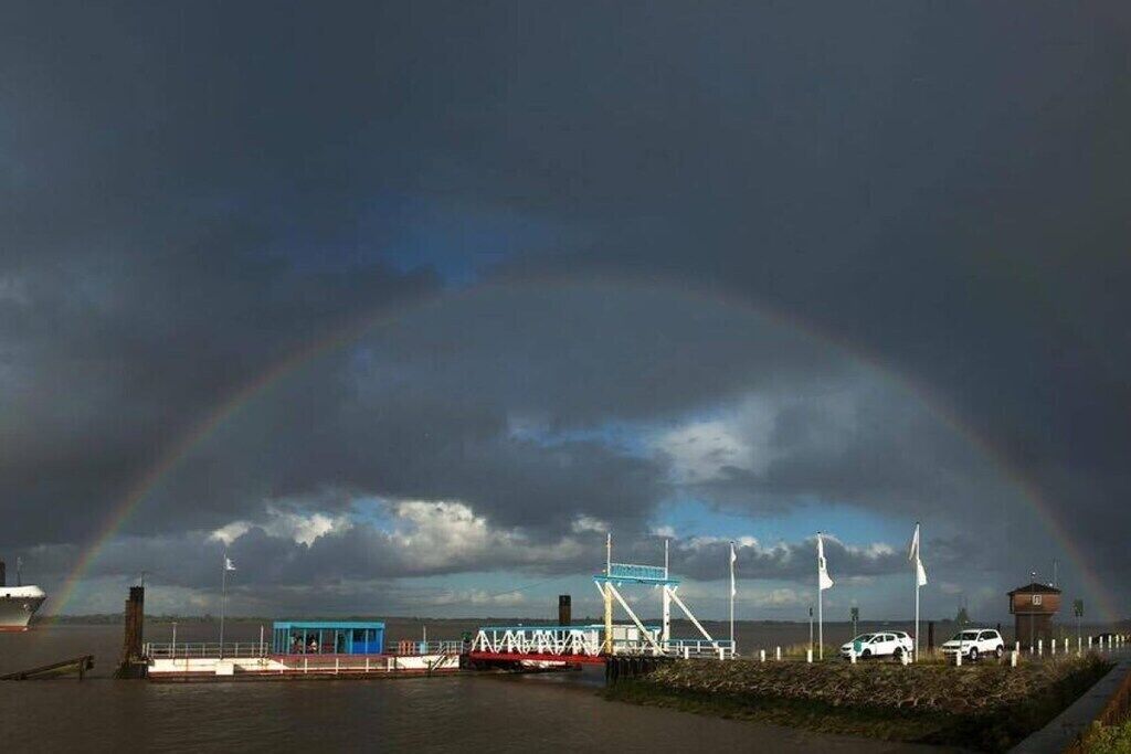 A coastal scene with a rainbow, a pier, a small blue building, and a flag-flagged structure.