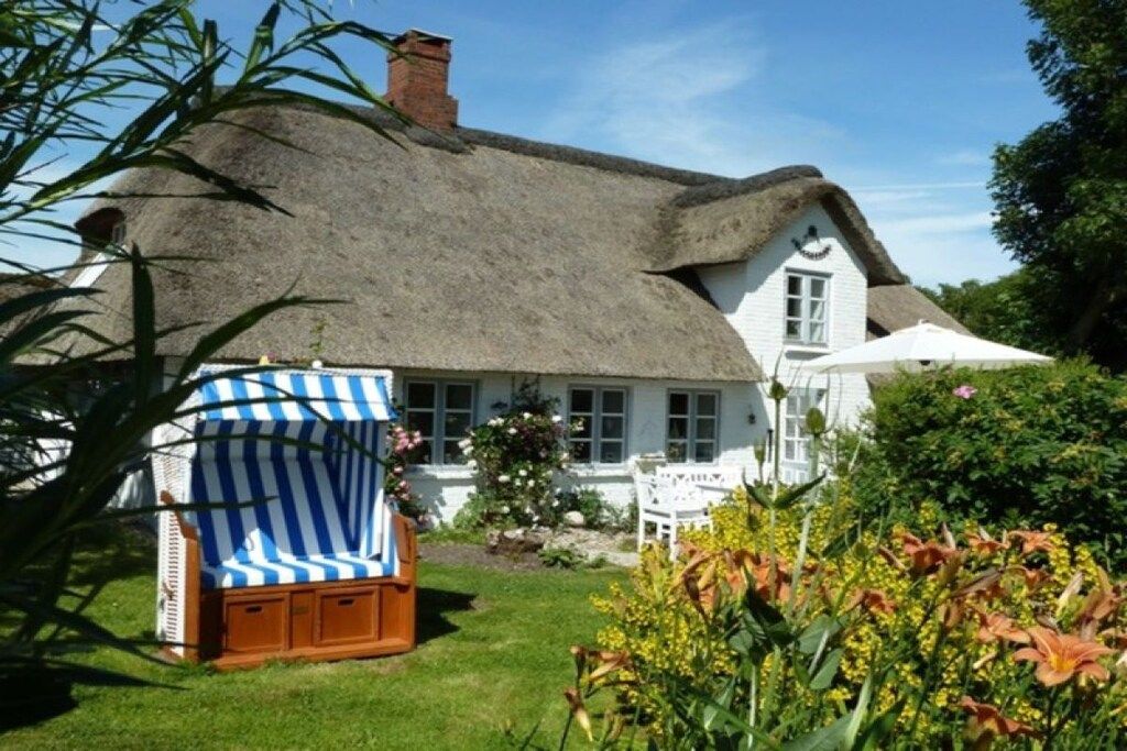 Un cottage con tetto di paglia e facciata bianca, un giardino con piante in fiore e una sedia da spiaggia a righe.
