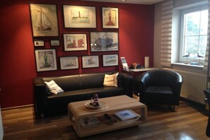 A room with a red wall, a black leather sofa, a wooden coffee table with a lighthouse model, and framed artwork. - Holiday apartment with 2 bedrooms (Norderney)