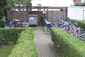 Ein gepflasterter Weg führt zu einem überdachten Fahrradstellplatz mit Holzzaun und Mülleimer.