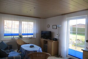 A room with a television, a table with a blue tablecloth, a wicker chair, a sofa, and a window with a view of a garden. - Holiday apartment with 1 bedroom (Groß Zicker)