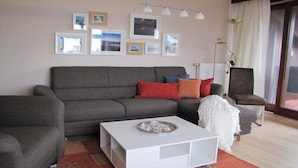 Una sala de estar con un sofá gris, una mesita de centro blanca y una silla. Hay cuadros en la pared y una lámpara.