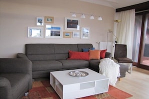 Ein Wohnzimmer mit grauem Sofa, einem weißen Couchtisch und einem Sessel. An der Wand hängen gerahmte Bilder und es steht eine Lampe bereit.