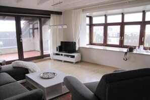 Un salon moderne avec un téléviseur à écran plat posé sur un meuble blanc, des canapés gris, une table basse et une grande fenêtre avec des rideaux.