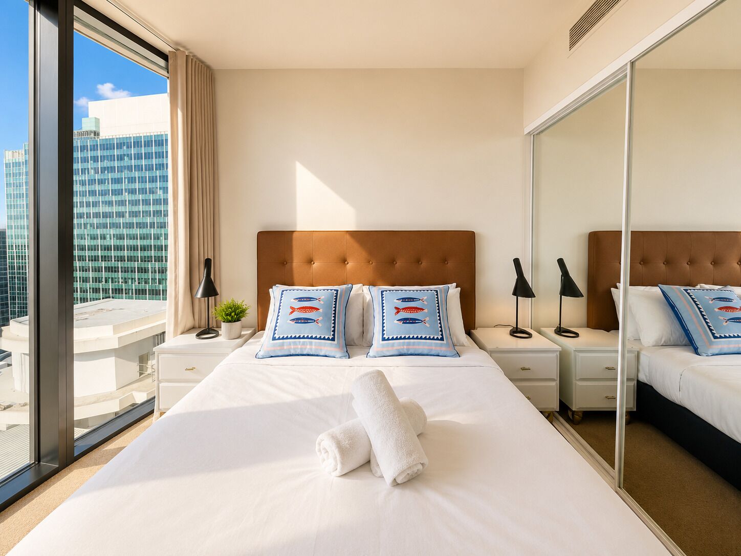 A hotel room with a large bed, two bedside tables, a nightstand, a mirror, and a view of a cityscape through large windows.