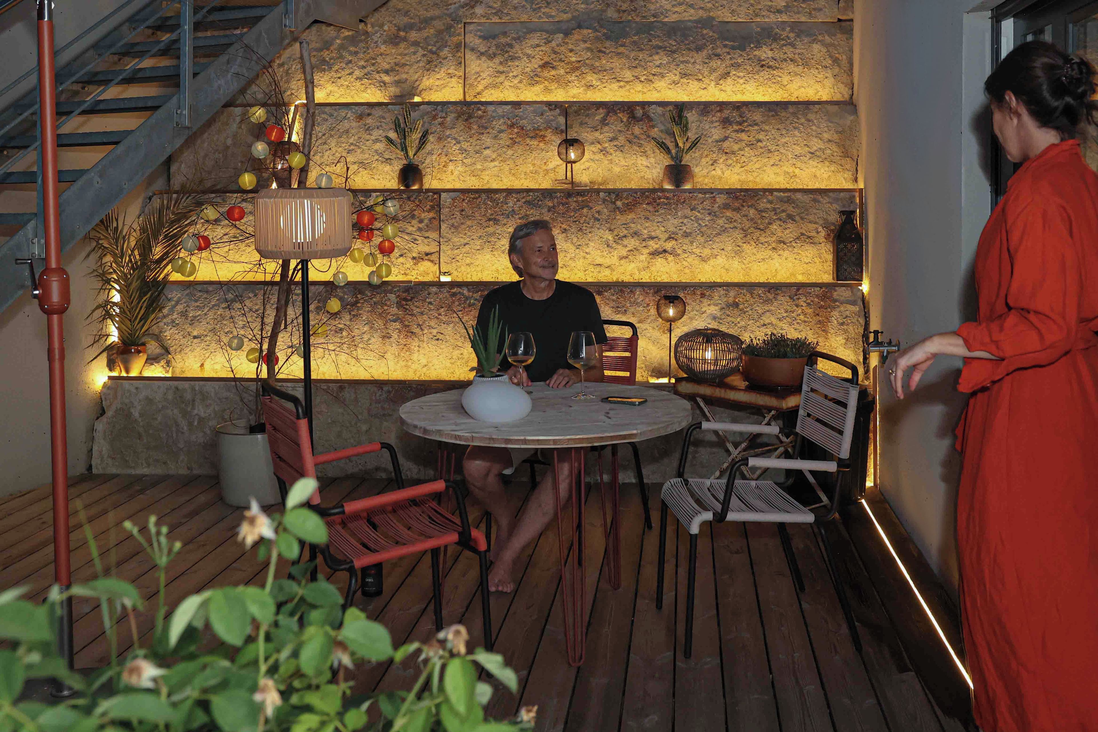 A man seated at a round table with a wine glass, surrounded by chairs and a rustic stone wall with decorative lights.