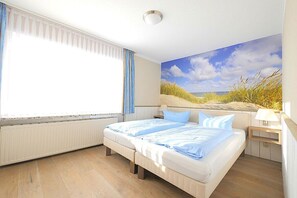 A bedroom with a large bed, a window with curtains, a wall mural, and a bedside table with a lamp. - Seestern 10 Apartment House Seestern (Norderney)