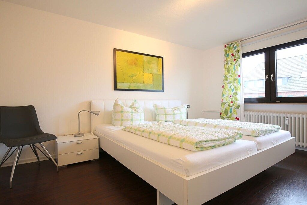 A bedroom with a bed, a chair, a nightstand, a lamp, a radiator, and a window with curtains.