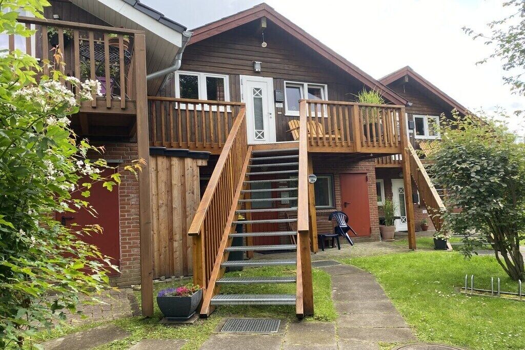 Una casa de madera de dos pisos con un balcón, una escalera y un pequeño jardín.
