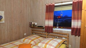 A wooden-paneled room with a bed, a window with curtains, a shelf with books and a lamp, and a wall-mounted clock. - Holiday apartment in Holnis with Wi-Fi (Holnis)