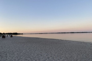 Una playa con un mar tranquilo, una orilla arenosa y algunas construcciones a lo lejos.