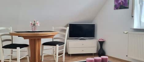 Ein Zimmer mit Esstisch, Stühlen, Fernseher, einer kleinen Kommode und einem Fenster mit einem Blumenbild.