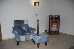 Ein Zimmer mit blauem Sessel, kleinem Tisch mit einem Buch, Stehlampe und Kaminofen.