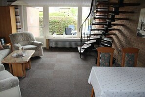 A room with a sofa, armchair, and a table with a vase. There is a window with a view of greenery outside. - Sailor House Semi-detached (Dornumersiel)
