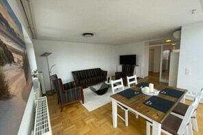 Ein Wohnzimmer mit einer Couch, Sesseln, einem Esstisch mit Platzsets und einem Fernseher.