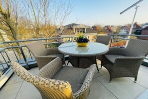 A round glass table with wicker chairs and a yellow flower pot on a balcony with a metal railing. - Holiday apartment with 2 bedrooms (Bad Zwischenahn)
