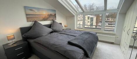 Un dormitorio con tragaluz, una cama con ropa de cama gris, una mesita de noche con lámpara y un cuadro en la pared.