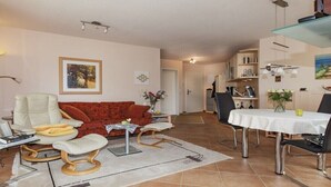 A living room with a sofa, a dining table with chairs, and a kitchen in the background. - Holiday apartment in Sylt with swimming pool (Sylt)