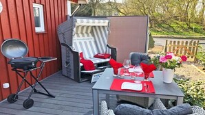 A deck with a grill, a chair with a striped cushion, a table set for dining, and a wicker chair with cushions. - Holiday home with 1 bedroom (Grödersby)