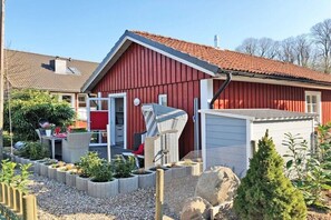 Ein rot gestrichenes Backsteingebäude im Stil eines Bauernhauses mit geneigtem Dach, einem von Zäunen umgebenen Garten mit verschiedenen Pflanzen und einem kleinen Schuppen.