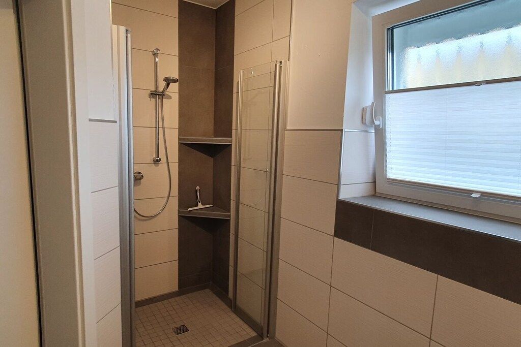 A small, enclosed shower area with a glass door, a window with blinds, and a tiled floor.