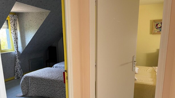 A bedroom with a bed, a chair, a radiator, and a window with curtains. - Normandy Gite With Bay Views (Courtils)