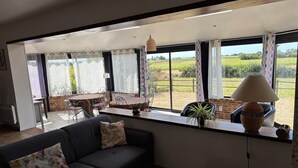 A living room with a sofa, a dining table, and chairs, a window with a view of a field, and a lamp on a side table. - Normandy Gite With Bay Views (Courtils)