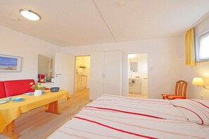 A hotel room with a bed, a dining table, a red chair, a small table with a vase of flowers, and a kitchen area visible through an open door. - Seestern 5 Apartment House Seestern (Norderney)