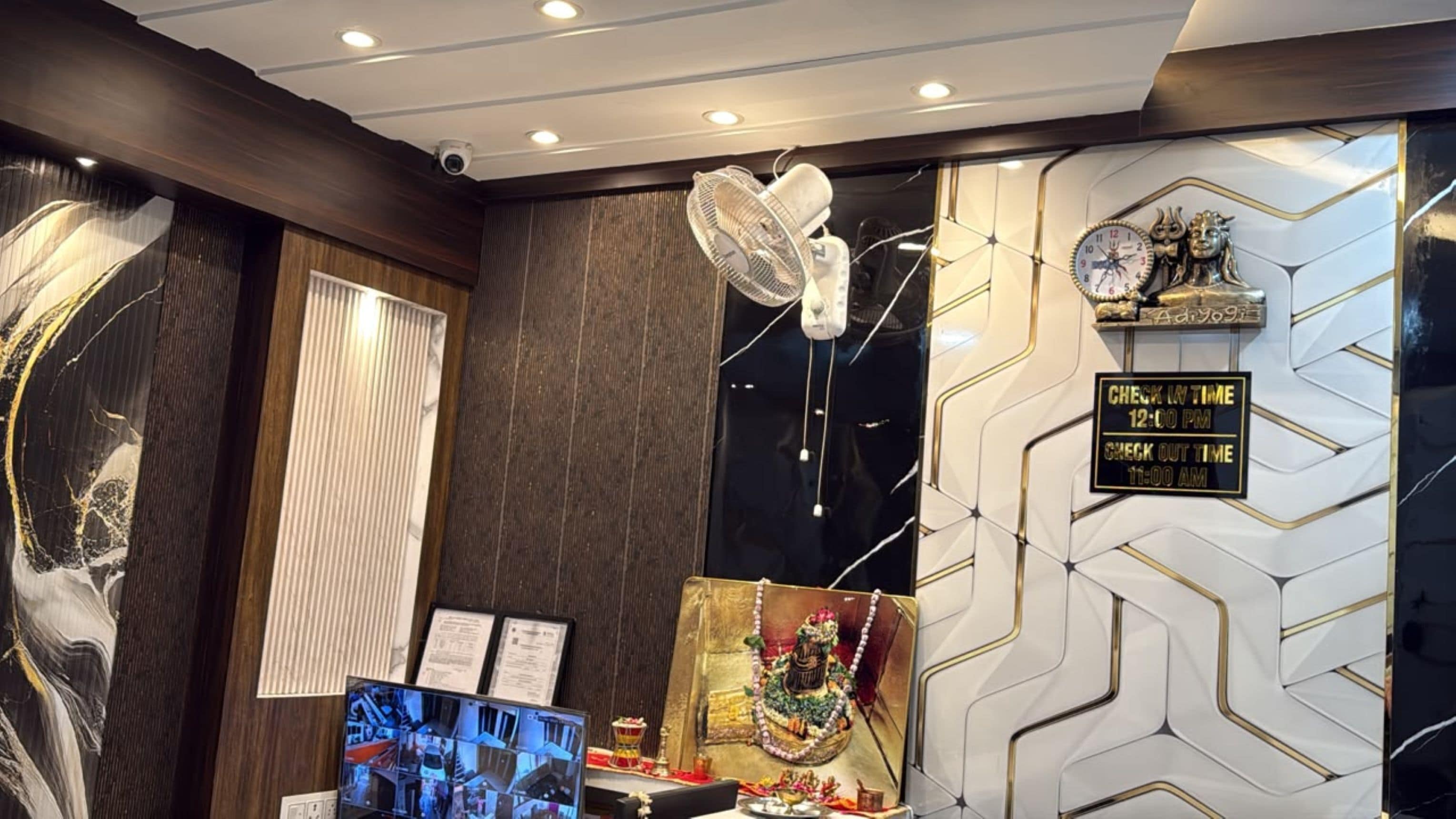 Uma área de recepção de hotel com balcão de check-in, câmera de segurança, relógio de parede e uma parede decorativa com padrão geométrico.