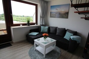 Ein modernes Wohnzimmer mit einer schwarzen Couch, einem weißen Couchtisch und Blick auf ein Gewässer durch ein Fenster.