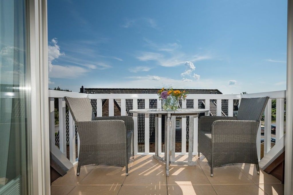A balcony with two wicker chairs and a small table, a white railing, and a clear sky.