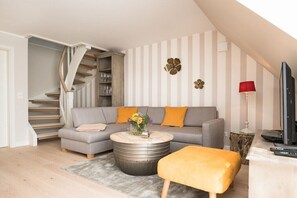 A modern living room with a staircase, a grey sofa with orange cushions, a round coffee table with a flower vase, and a red lamp. - Villa Jansen 5 - OG/DG (Wenningstedt)