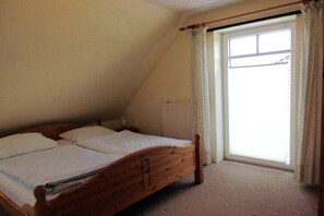 Un lit simple avec un cadre en bois, dans une pièce dotée d’un plafond en pente, d’une fenêtre avec des rideaux et d’un radiateur.