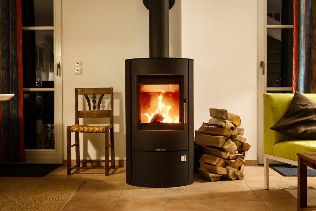 A modern living room with a wood-burning stove, a wooden chair, a sofa, and a stack of firewood.