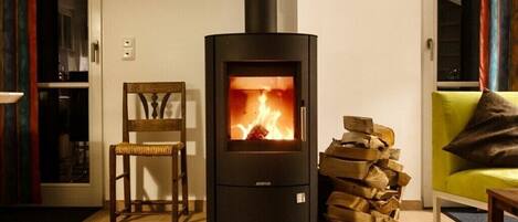 A modern living room with a wood-burning stove, a wooden chair, a sofa, and a stack of firewood.