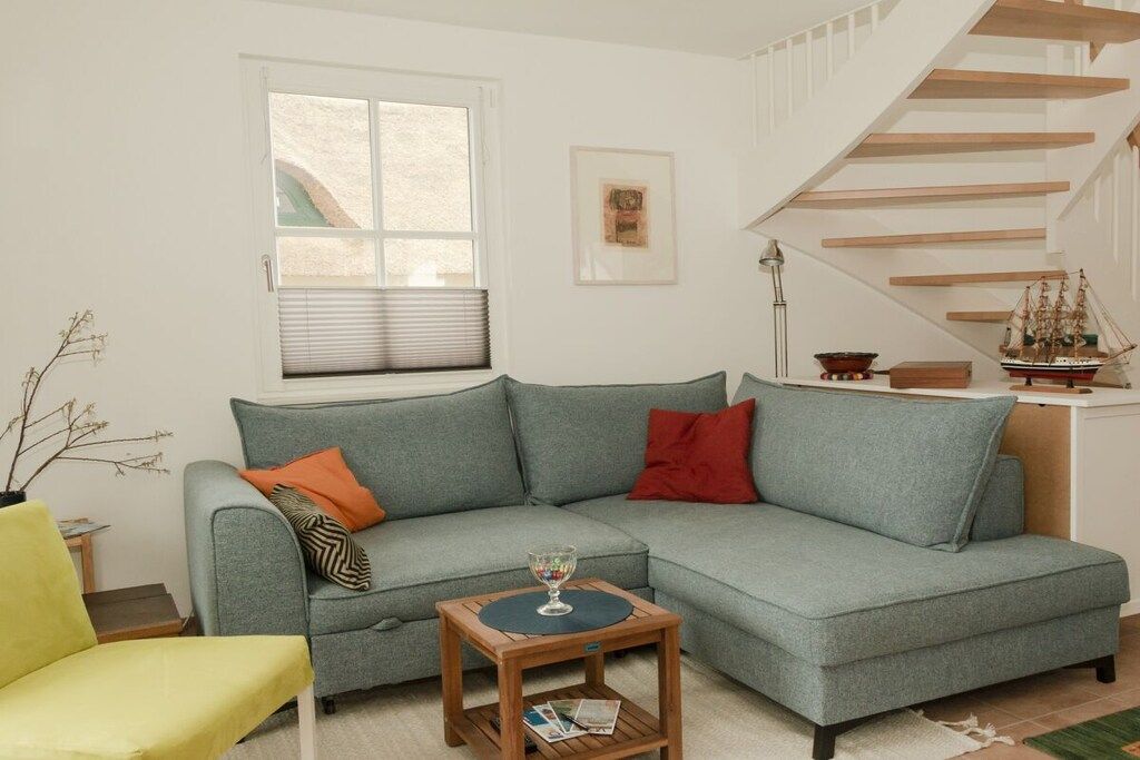 Ein Wohnzimmer mit einem hellblauen Sofa, einem Holztisch, einem gelben Sessel, einem Fenster mit Jalousien, einem gerahmten Bild an der Wand und einer Treppe mit einem Schiffmodell auf der Setzebene.