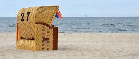 Een rieten strandstoel met een vlag, nummer 27, op een zandstrand met een rustige zee en een schip in de verte.