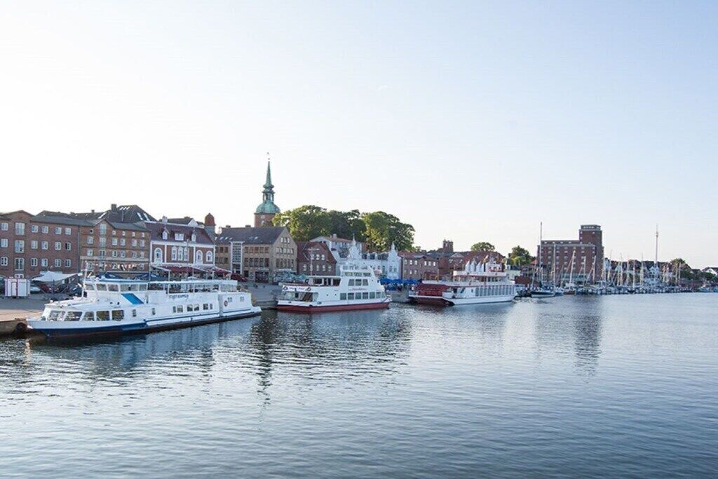 A waterfront with several boats docked, a prominent church spire, and a row of buildings along the shore.